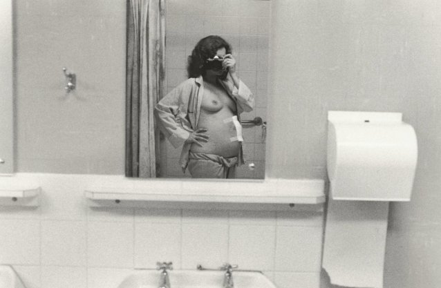 3/4 length self portrait, camera held to face, pyjama top open, 1979