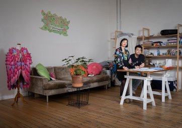 Two people at a bench covered in sheets of paper in a room with a wooden floor and a large brown couch