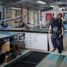 Alison Alder wearing a paint splattered apron, leaning against a bench in her studio