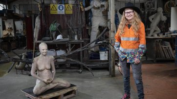 Nell standing in her workshop next to a cross legged sculpture